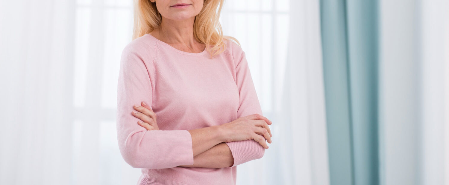 Mulher madura de cabelos loiros e blusa rosa com os braços cruzados em ambiente claro.