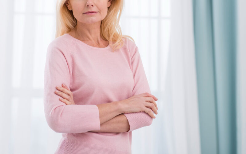 Mulher madura de cabelos loiros e blusa rosa com os braços cruzados em ambiente claro.